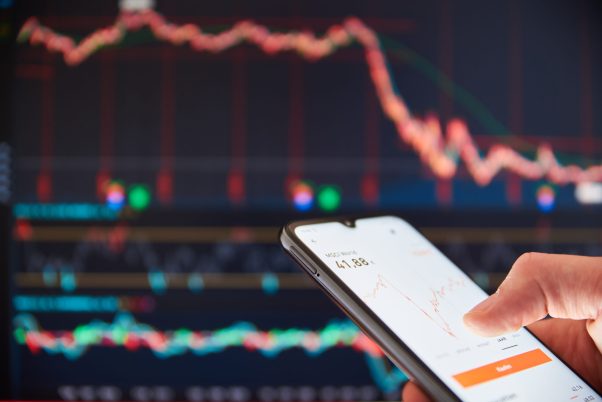Person checking the stock prices on a smartphone in front of a big screen market rates. Person viewing share prices on a smartphone with market prices on a screen in the background.